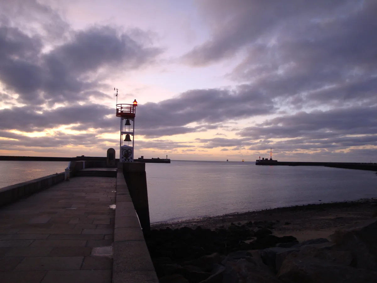 A pier with a red bell beacon leading out to the harbour entrance at Le Havre under a dramatic sunset sky.