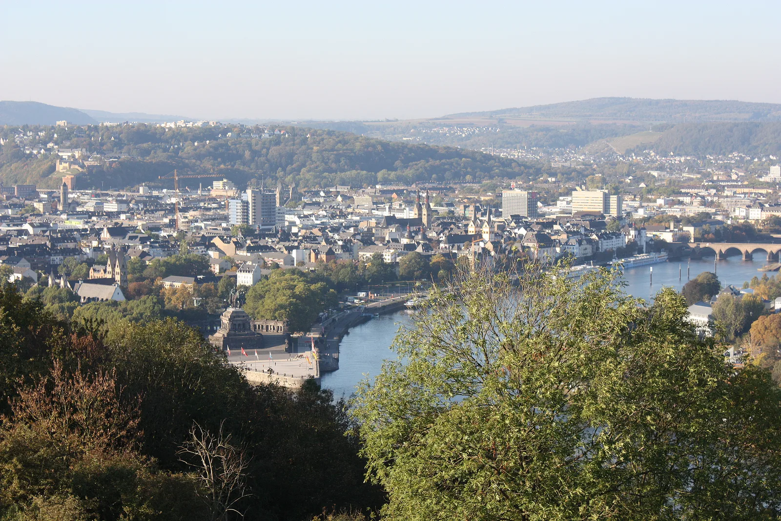 A wide view over Koblenz rooftops and the Moselle from the Ehrenbreitstein fortress hill under a pale sky.